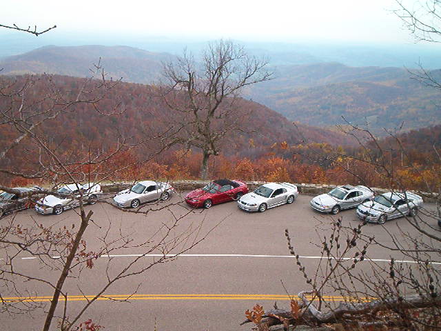 Several cars parked on a scenic overlook with autumn foliage in the background