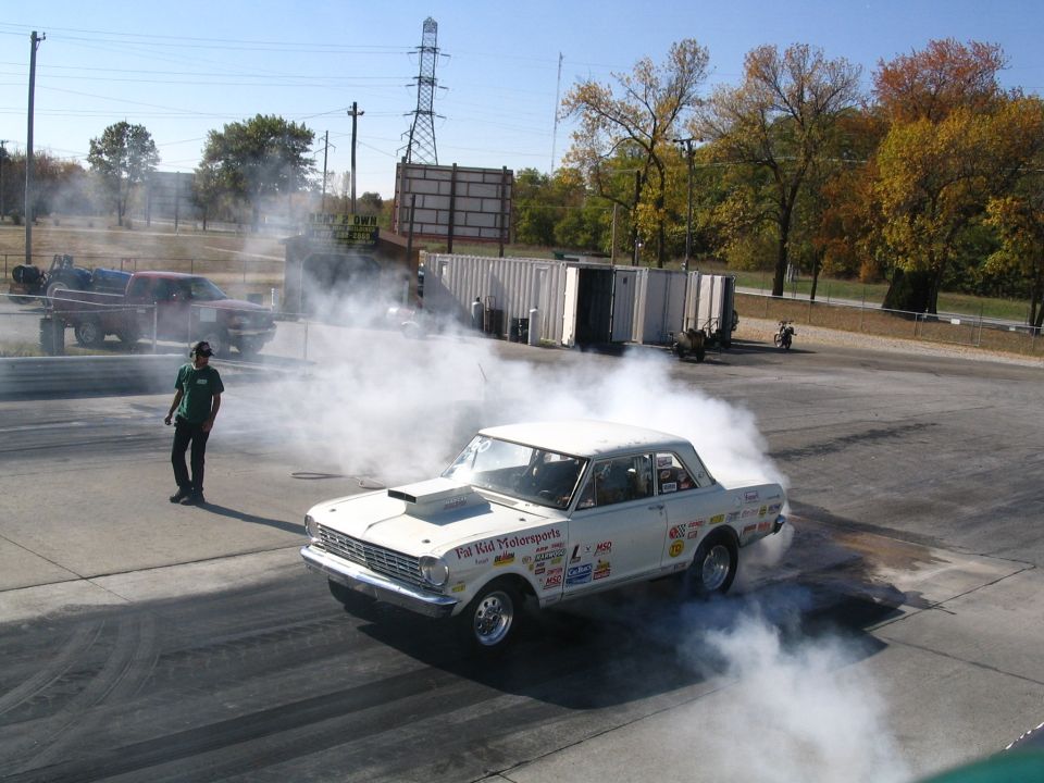 White Chevy Nova drag car with hood scoop doing a burnout, smoke behind it, at a drag strip