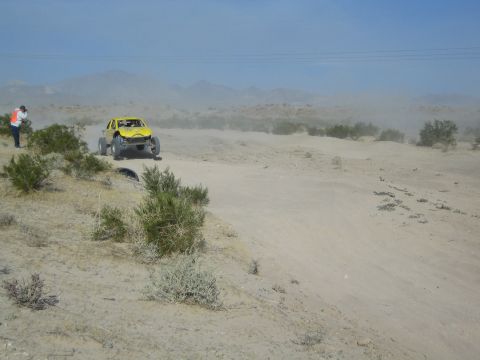 Yellow off-road buggy climbing a sandy berm past a corner worker in orange vest