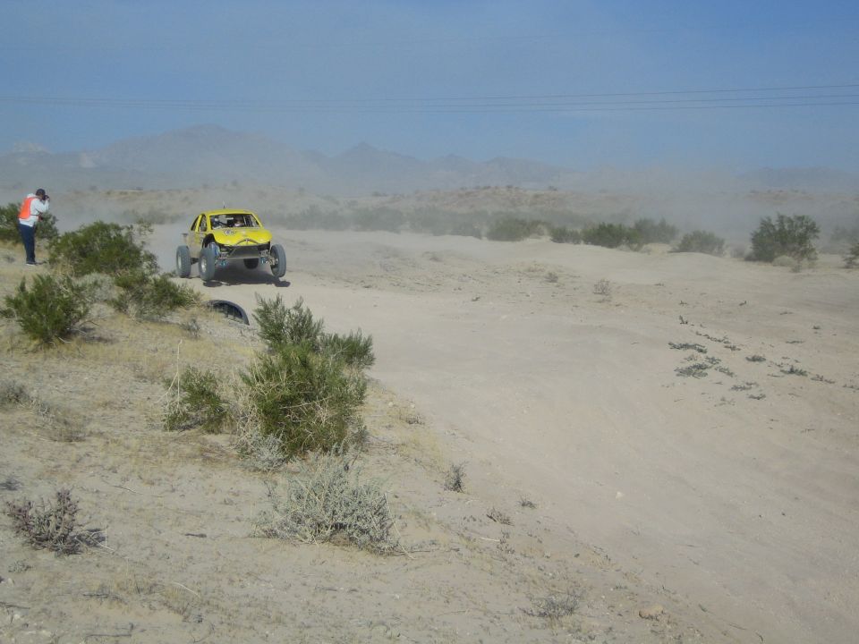 Yellow off-road buggy climbing a sandy berm past a corner worker in orange vest