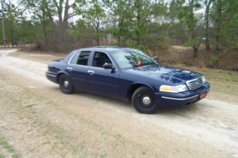 1998 Ford Crown Victoria - This dark blue sedan is parked on a dirt road with trees in the background.
