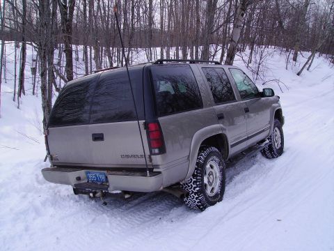 2000 Chevrolet Tahoe - A silver Chevrolet Suburban navigates a snowy path, its tires equipped with snow chains for traction.