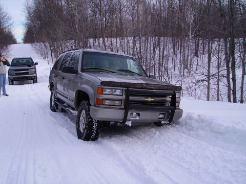 2000 Chevrolet Tahoe - A silver SUV with a bull bar is stuck in the snow, with another vehicle visible behind it on a wooded road.