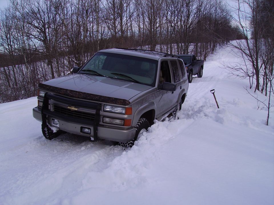Silver Chevrolet Tahoe stuck in deep snow on a wooded road, with a black pickup truck behind it. A shovel is stuck in the snow