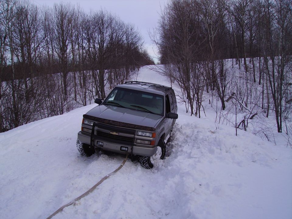 Gray Chevrolet Tahoe stuck in deep snow on a wooded road, with a tow strap attached to the front