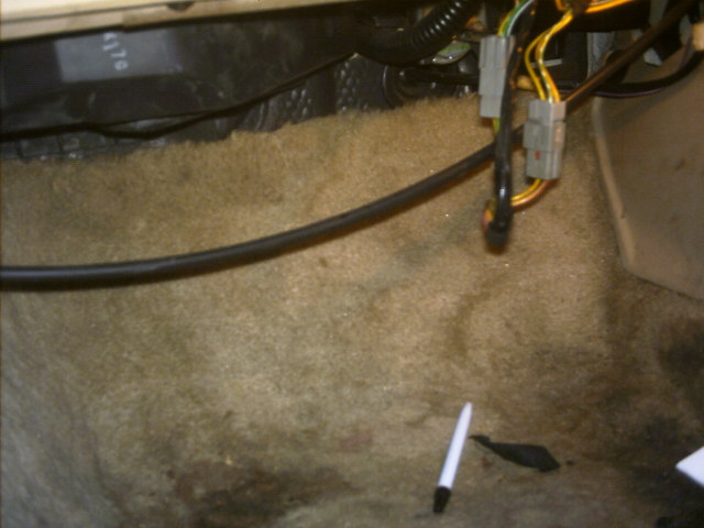 Car interior floor with tan carpet, black wiring harness, and a white pen