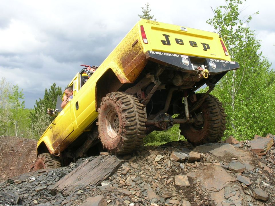 Yellow Jeep pickup crawling up a rocky slope, rear view showing lifted suspension and muddy off-road tires