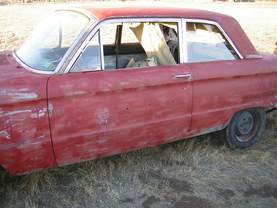 1963 Ford Falcon sedan side view, red paint faded and showing primer, door handle present, interior covered with tarp