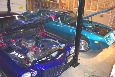 1979 Pontiac Trans Am - Two muscle cars, a purple one and a teal one, are parked in a garage with their hoods open, revealing their engines.