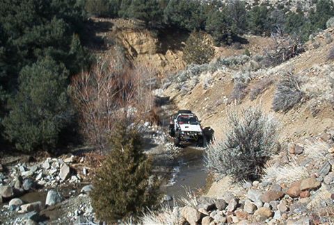 1986 Ford Bronco - A white off-road vehicle drives through a shallow stream in a , natural landscape.