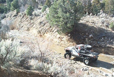 1986 Ford Bronco - Custom off-road truck navigates a rocky trail in a dry, brushy landscape.