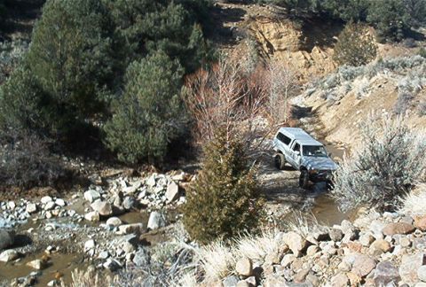 1986 Ford Bronco - A silver SUV navigates a rocky, dry stream bed surrounded by arid hills and sparse trees.