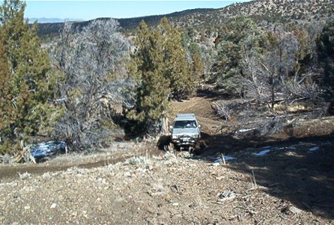 1986 Ford Bronco - A silver SUV navigates a muddy dirt road through a sparse, wooded landscape under a clear blue sky.
