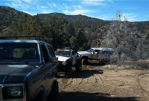 1986 Ford Bronco - Three off-road vehicles are parked on a dirt track in a hilly, wooded area.