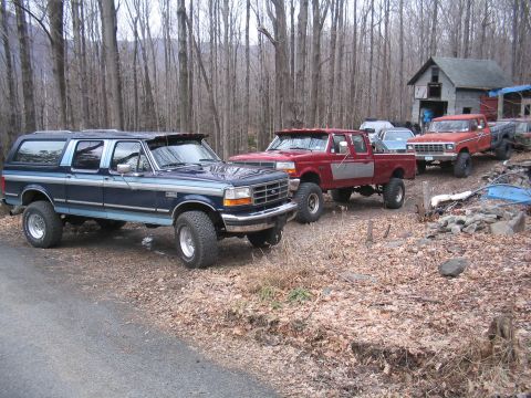1979 Ford Bronco - A lifted blue Ford Bronco SUV is parked in a wooded area alongside several pickup trucks.
