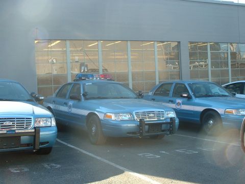 1999 Ford Crown Victoria - Multiple light blue police cars are parked in front of a large garage with many windows.
