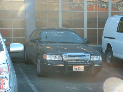 1999 Ford Crown Victoria - A black Ford Crown Victoria police car is parked in a garage next to a white van.