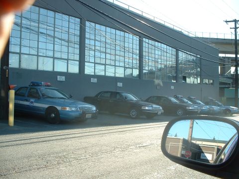 1999 Ford Crown Victoria - Several police sedans and other dark-colored sedans are parked in a row outside a large industrial building.