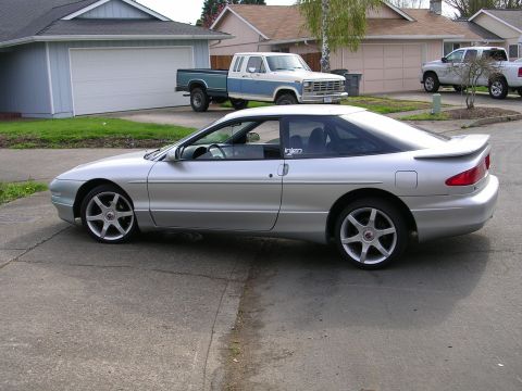 1996 Ford Probe - A silver 1996 Ford Probe coupe is parked in a driveway, with two pickup trucks visible in the background.