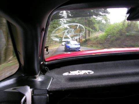 1996 Ford Probe - A blue sports car is seen driving on a road surrounded by trees, viewed through the rear window of another car.