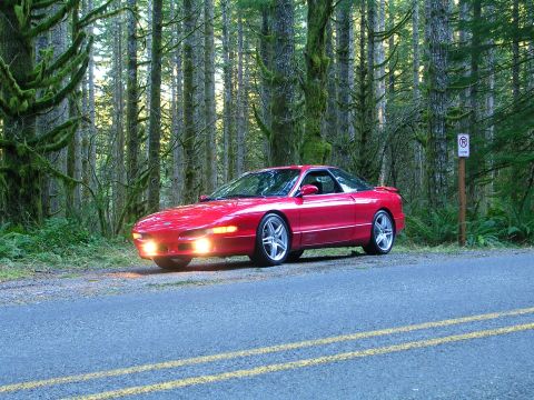 1996 Ford Probe - A red 1996 Ford Probe is parked on the shoulder of a road surrounded by a dense forest.
