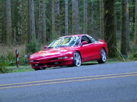 1996 Ford Probe - A red 1996 Ford Probe sits parked on the shoulder of a road, surrounded by tall trees.