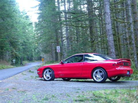 1996 Ford Probe - A red 1996 Ford Probe GT sits on a gravel shoulder beside a winding road through a moss-covered forest.