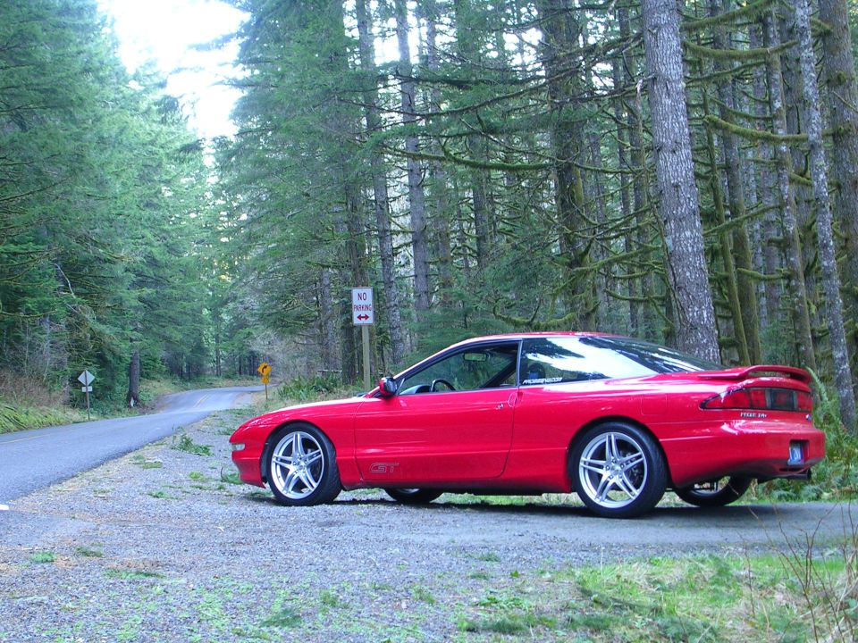Red Ford Probe GT coupe parked on gravel beside a forest road, side view