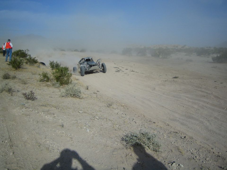 Open-frame off-road buggy racing along a desert trail with a flagger watching