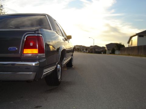 1988 Ford Crown Victoria LTD - The rear of a dark sedan is visible from a low angle on a residential street at dusk.