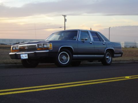 1988 Ford Crown Victoria LTD - A 1988 Ford Crown Victoria LTD is seen from the front-three-quarter view on a road with yellow double lines.