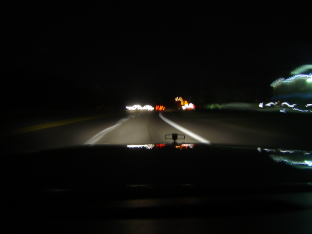 View from inside a car at night, looking at the dashboard and road ahead with headlights illuminating the way