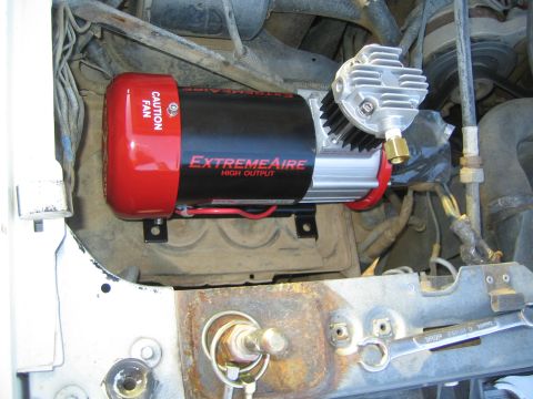1990 Ford Bronco - red and silver ExtremeAire High Output air compressor with a CAUTION FAN cover sits mounted in a dusty engine bay.