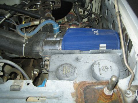 1990 Ford Bronco - blue cylindrical canister sits on a cast aluminum bracket among hoses and wiring in a dusty engine bay.