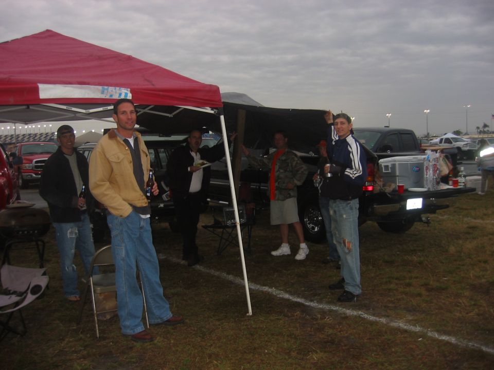 Tailgaters under a red canopy in a grass parking lot at dusk with trucks behind