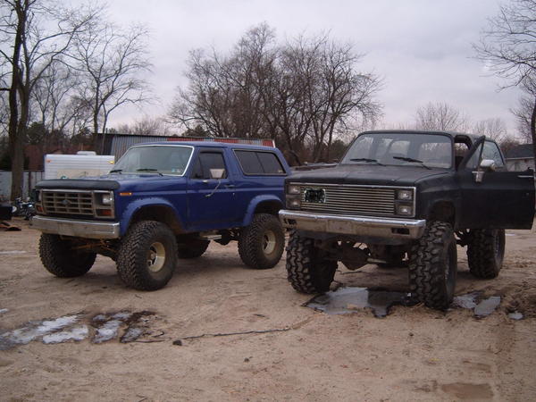 Lifted blue Ford Bronco on large off-road tires parked beside a black lifted SUV in a dirt lot