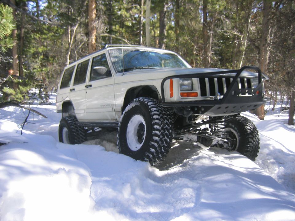 White 1997 Jeep Cherokee with large off-road tires driving through snow, a custom front bumper