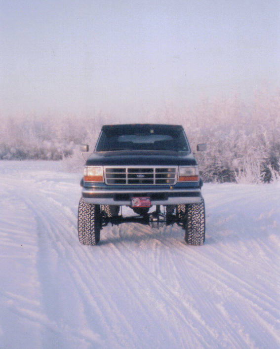 Dark lifted Ford truck facing camera on snowy road with frosty trees behind