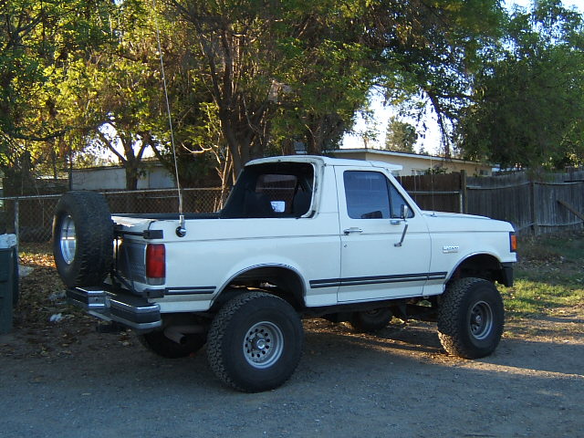 White Ford Bronco, lifted, with spare tire mounted on rear carrier. Side view shows aftermarket wheels and tires
