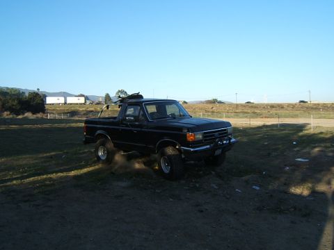 1988 Ford Bronco - Black Ford Bronco with a roof rack is parked on a dirt area, kicking up dust.