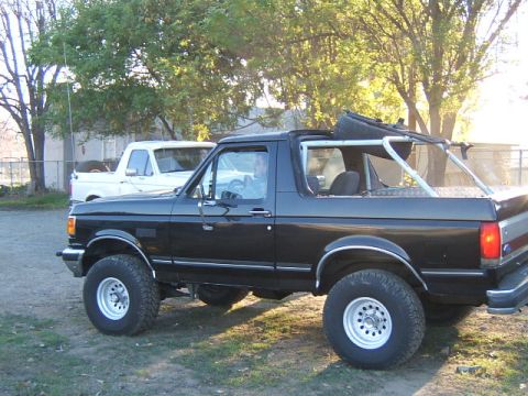 1988 Ford Bronco - Black full-size Ford Bronco with the top off, lifted on chrome-lip white wheels, parked beside a white pickup.
