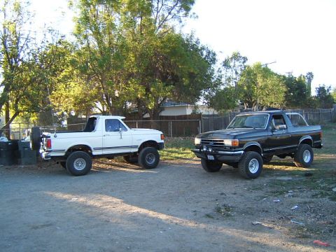 1988 Ford Bronco - Two Ford Broncos, a white one and a black one, are parked side-by-side on a dirt lot.