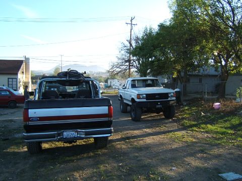1988 Ford Bronco - Two Ford pickup trucks are parked on a dirt road in front of houses and trees.