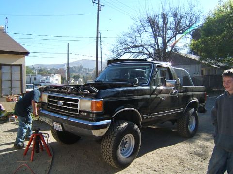 1988 Ford Bronco - A person works on the engine of a black Ford Bronco parked outside a house on a sunny day.