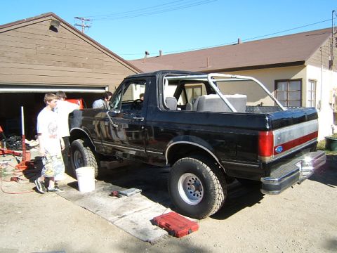1988 Ford Bronco - A black Ford Bronco is parked outside a garage with a roll cage installed and people working on it.