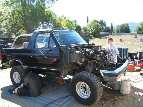 1988 Ford Bronco - A black Ford Bronco is undergoing mechanical work outdoors, with its hood and front end disassembled.