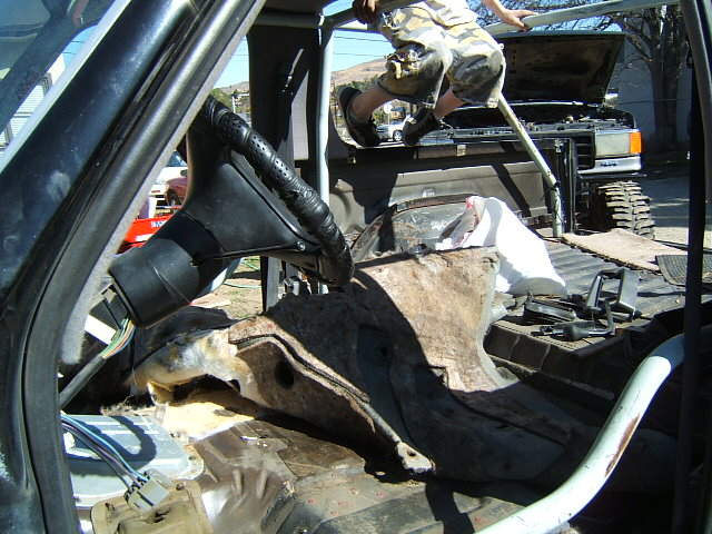 Disassembled vehicle interior with exposed wiring and rusted floor panels. A person is visible climbing over the rear