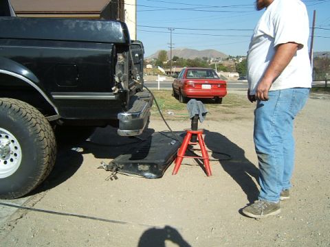 1988 Ford Bronco - A black pickup truck's fuel tank is removed and resting on the ground, with a red sedan parked nearby.