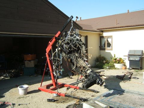 1988 Ford Bronco - An engine and transmission assembly is suspended by a red engine hoist in front of a garage and house.
