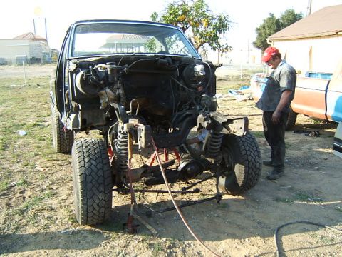 1988 Ford Bronco - A man works on a heavily dismantled truck chassis, its front end elevated on stands.